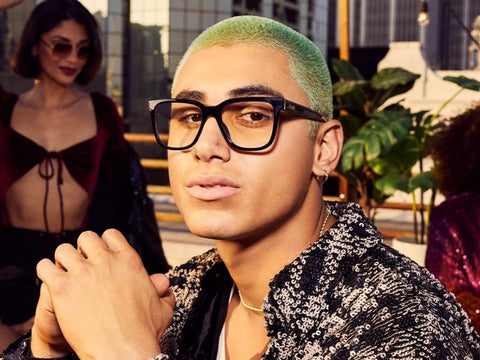 A male model posing with QUAY black prescription glasses with a female model in the background overlooking a city skyline